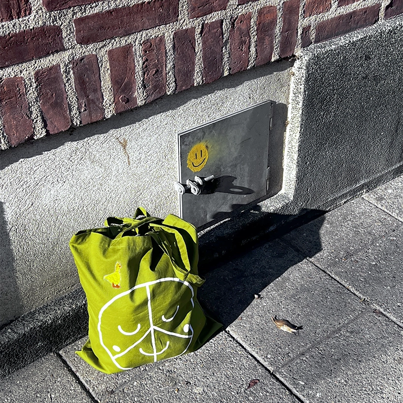 Green textile bag with a small embroidered duck, placed outdoors against a brick wall in sunlight